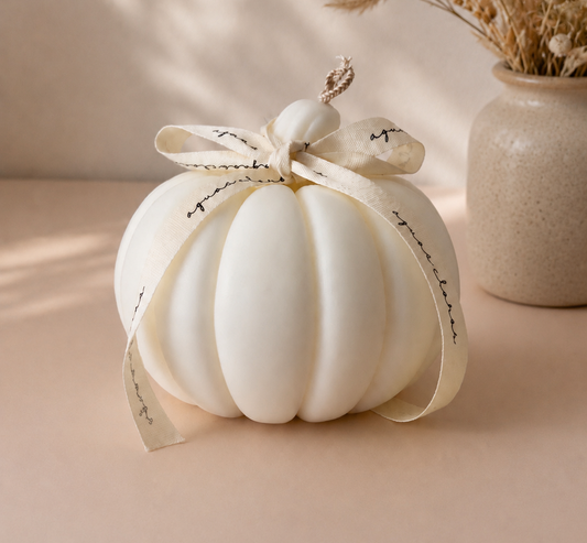 A small ribbed decorative pumpkin in warm off-white, tied with a cream ribbon printed with delicate black script, set on a beige surface with a soft-focus vase of dried grass in the background.