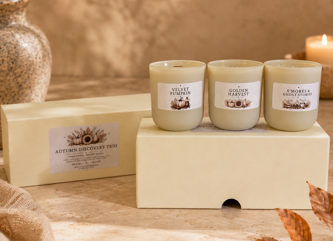 Three cream-colored scented candles in glass jars sit on a white display block next to their matching cream packaging box. The scene is styled with autumn leaves, a textured vase, and a warm candle in the background.