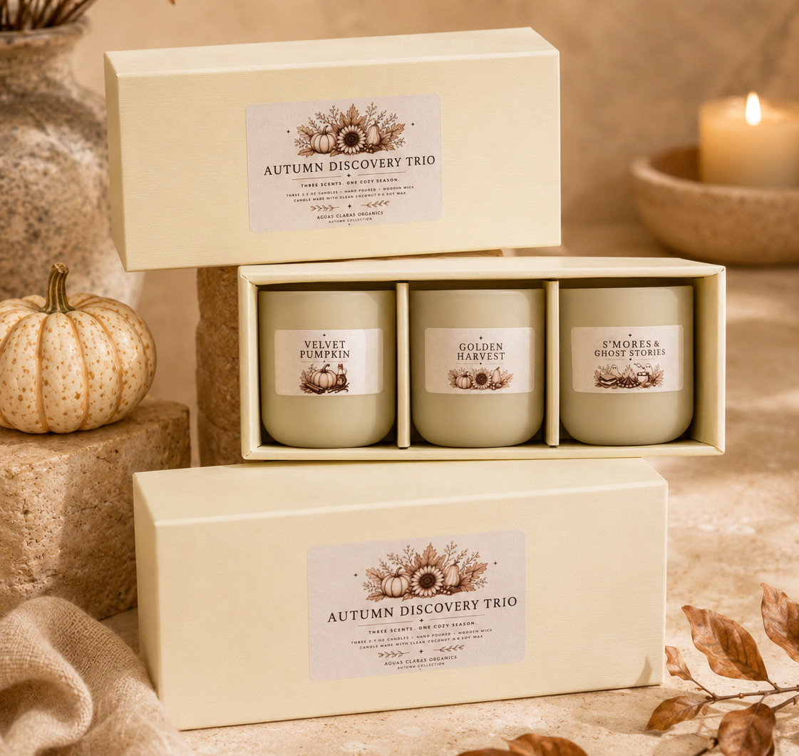 Three soy candles in a cream-colored gift box set labeled 'Autumn Discovery Trio' with scents Velvet Pumpkin, Golden Harvest, and Embrace the Fall, styled with autumn props like a small pumpkin, dried leaves, and a lit candle in the background.