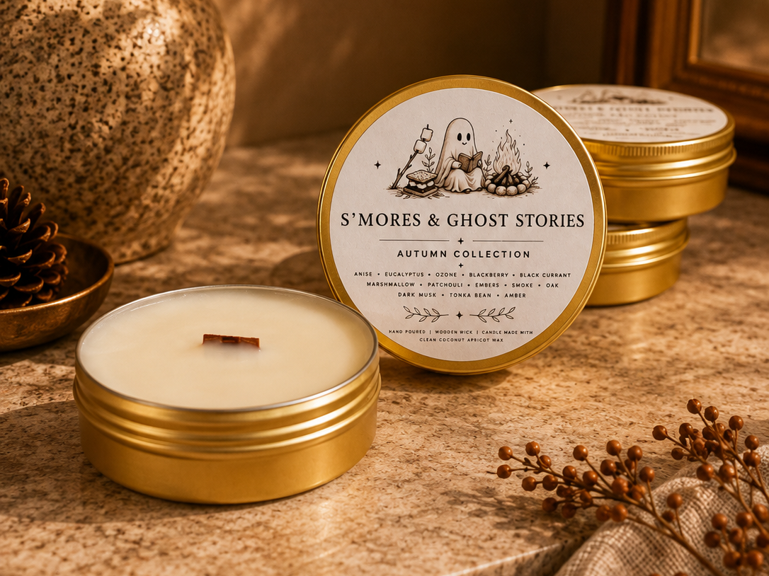 A round gold tin candle labeled 'S'MORES & GHOST STORIES' sits on a stone surface with another open candle in front and stacked tins behind. The scene includes a speckled vase with autumn leaves, a pinecone, and dried stems in a warm, styled setting.