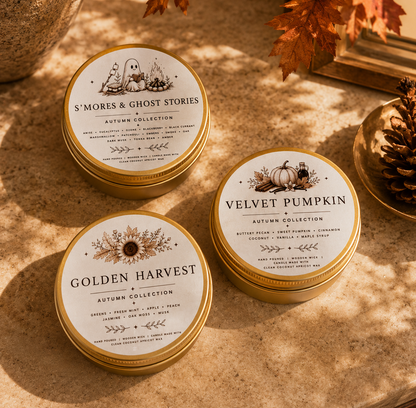 Three round metal tin candles from the Autumn Collection are arranged on a stone surface with fall-themed props. The tins are labeled Golden Harvest, Velvet Pumpkin, and S'mores & Ghost Stories, with gold lids and cream labels featuring autumn illustrations.