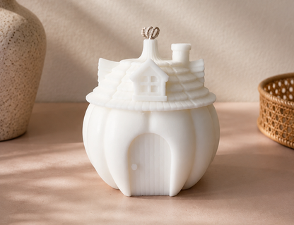 A white ceramic house-shaped candle holder with a small door and chimney detail, styled on a beige surface with soft shadows, a vase with dried stems on the left, and a woven basket on the right.